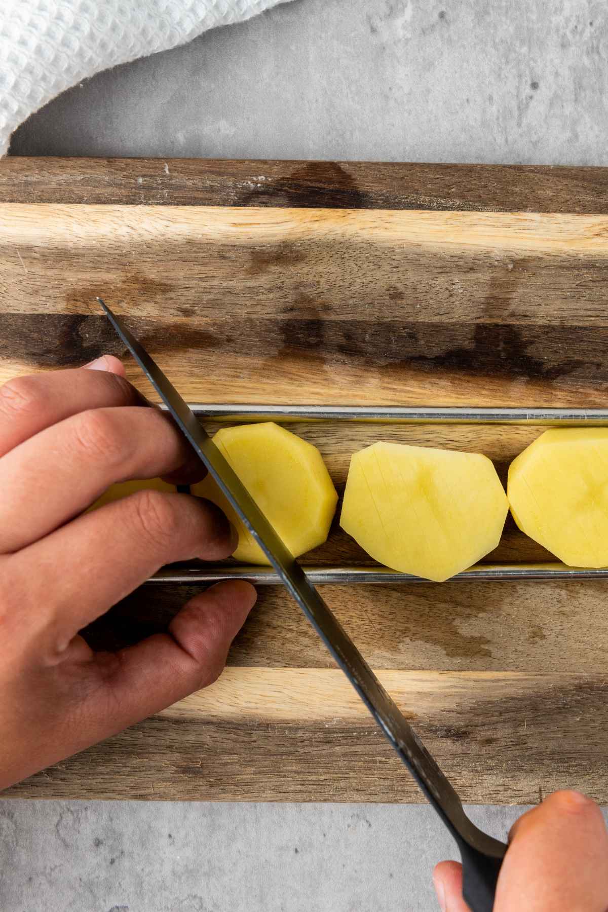 Close-up of a hand slicing a peeled potato placed between two skewers on a wooden cutting board to create accordion-style cuts.