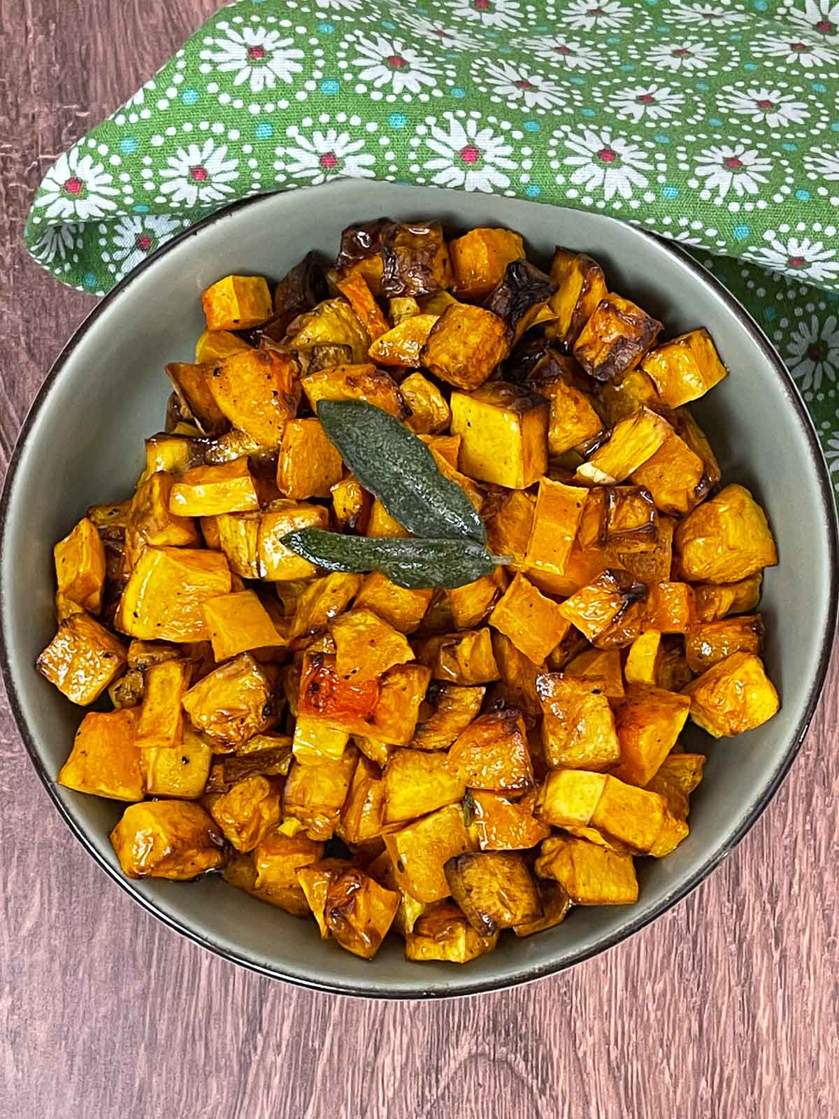 Overhead shot of a bowl of air fried butternut squash cubes with crispy edges, garnished with fried sage, set on a wood surface with a green floral cloth in the background.