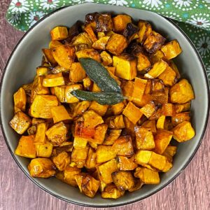 Overhead shot of a bowl of air fried butternut squash cubes with crispy edges, garnished with fried sage, set on a wood surface with a green floral cloth in the background.