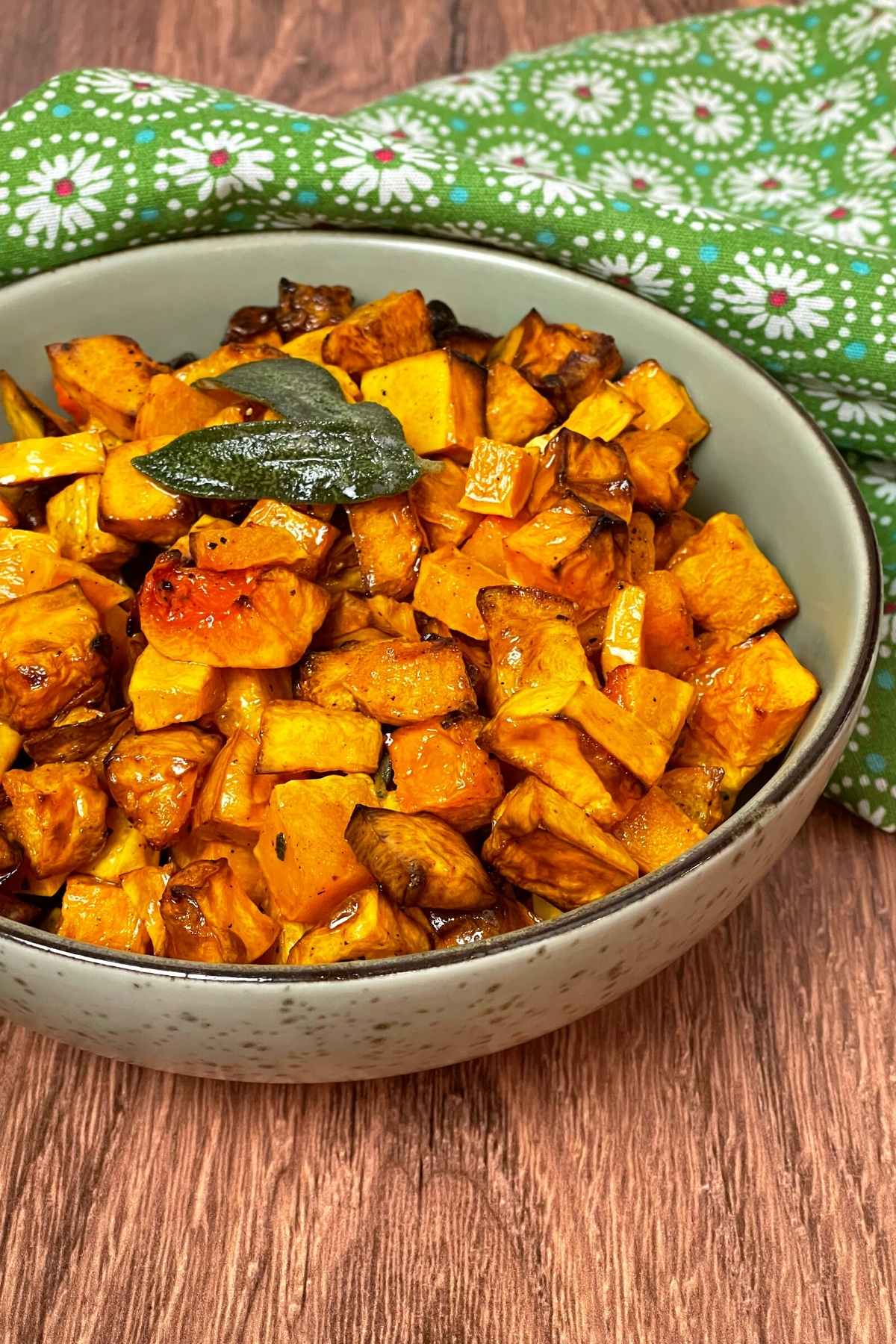 A bowl of roasted butternut squash cubes with crispy edges, garnished with fried sage, set on a wood surface with a green floral cloth in the background.