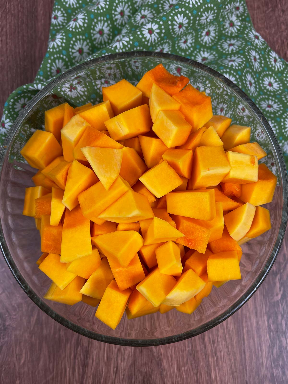 A glass bowl filled with freshly chopped raw butternut squash cubes, sitting on a wooden surface with a green and white floral cloth in the background.