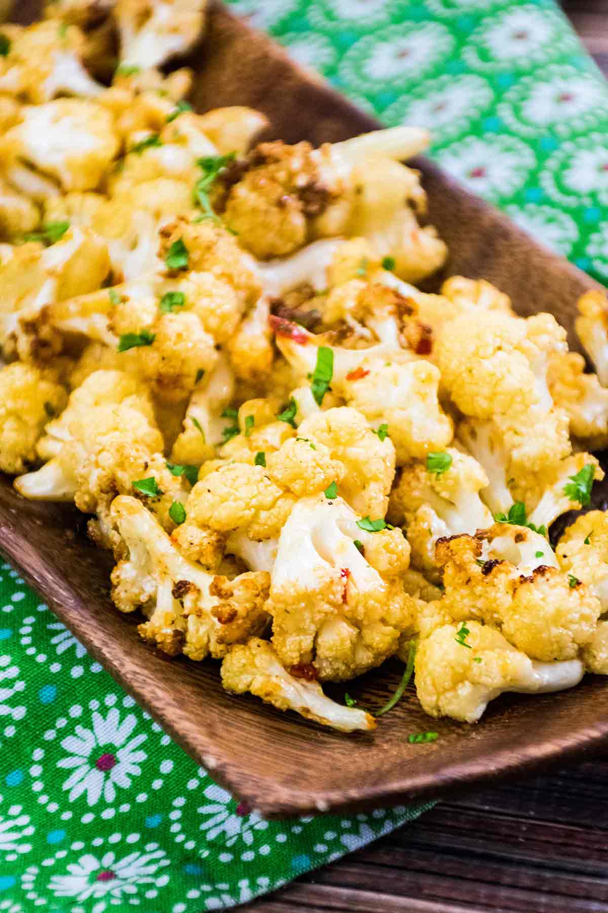 Close up of golden brown roasted cauliflower florets garnished with chopped cilantro, served on a wooden tray placed over a green floral-patterned cloth.