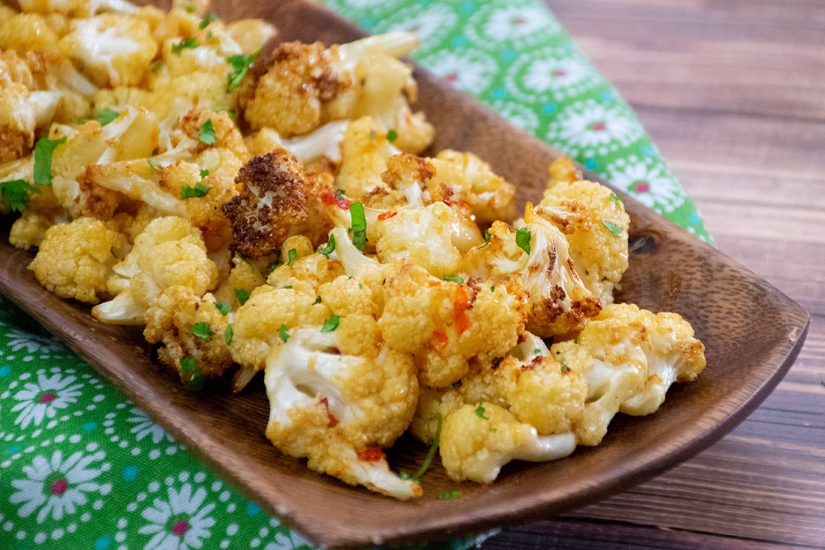Close up of golden brown roasted cauliflower florets garnished with chopped cilantro, served on a wooden tray placed over a green floral-patterned cloth.