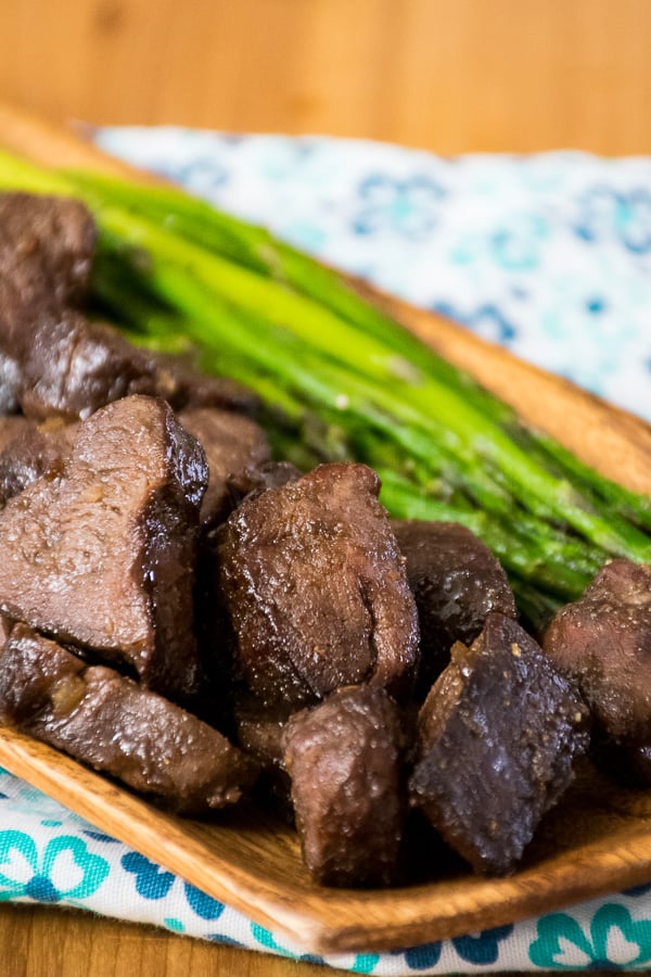 Air Fryer Venison Steak Bites plated on a wooden dish with a side of bright green asparagus spears on a blue floral napkin.