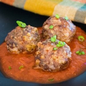 Three browned air fryer venison meatballs topped with chopped green onions, served in a pool of rich red tomato sauce on a dark plate with a colorful cloth napkin in the background.