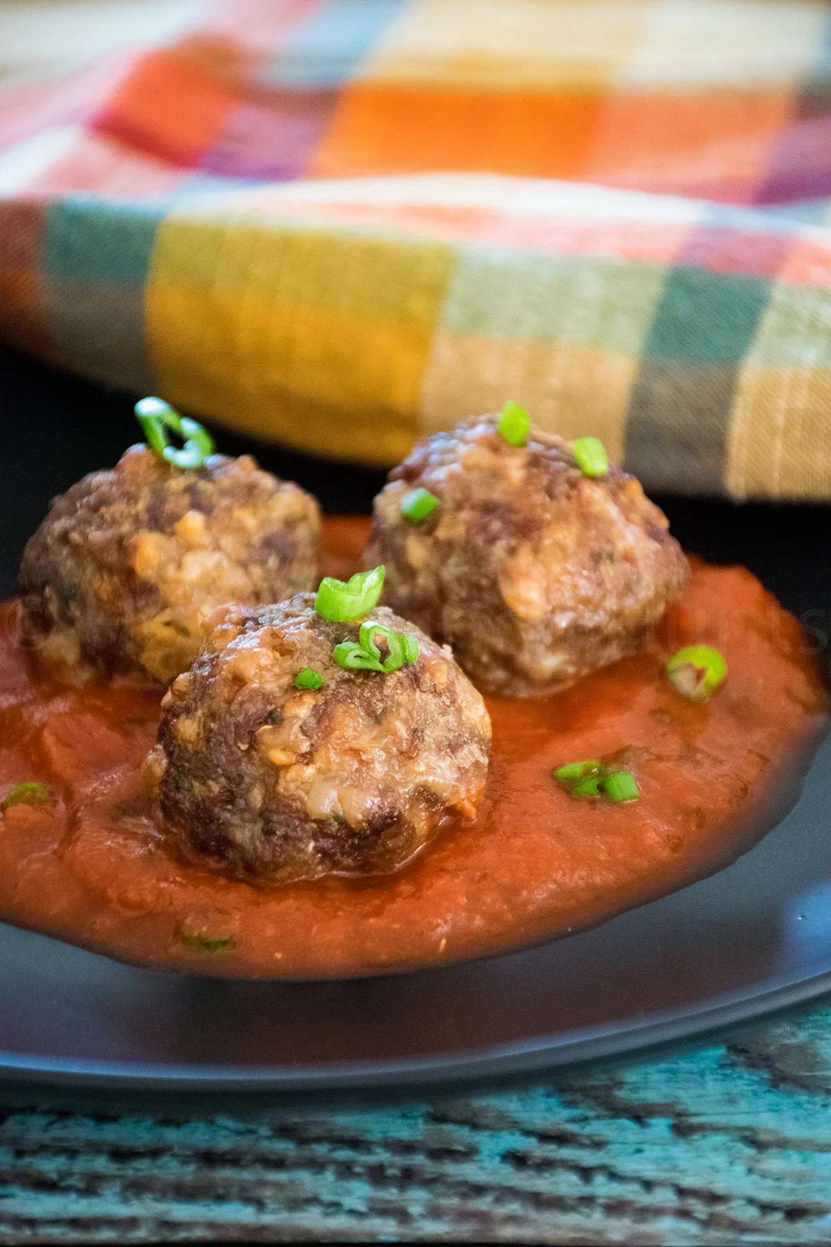 Three browned air fryer venison meatballs topped with chopped green onions, served in a pool of rich red tomato sauce on a dark plate with a colorful cloth napkin in the background.