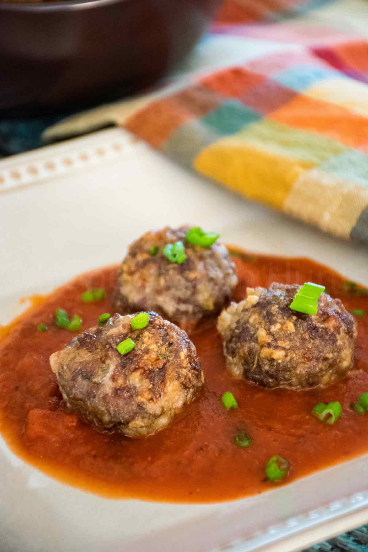 Three browned venison meatballs topped with chopped green onions, served in a pool of rich red tomato sauce on a dark plate with a colorful cloth napkin in the background.