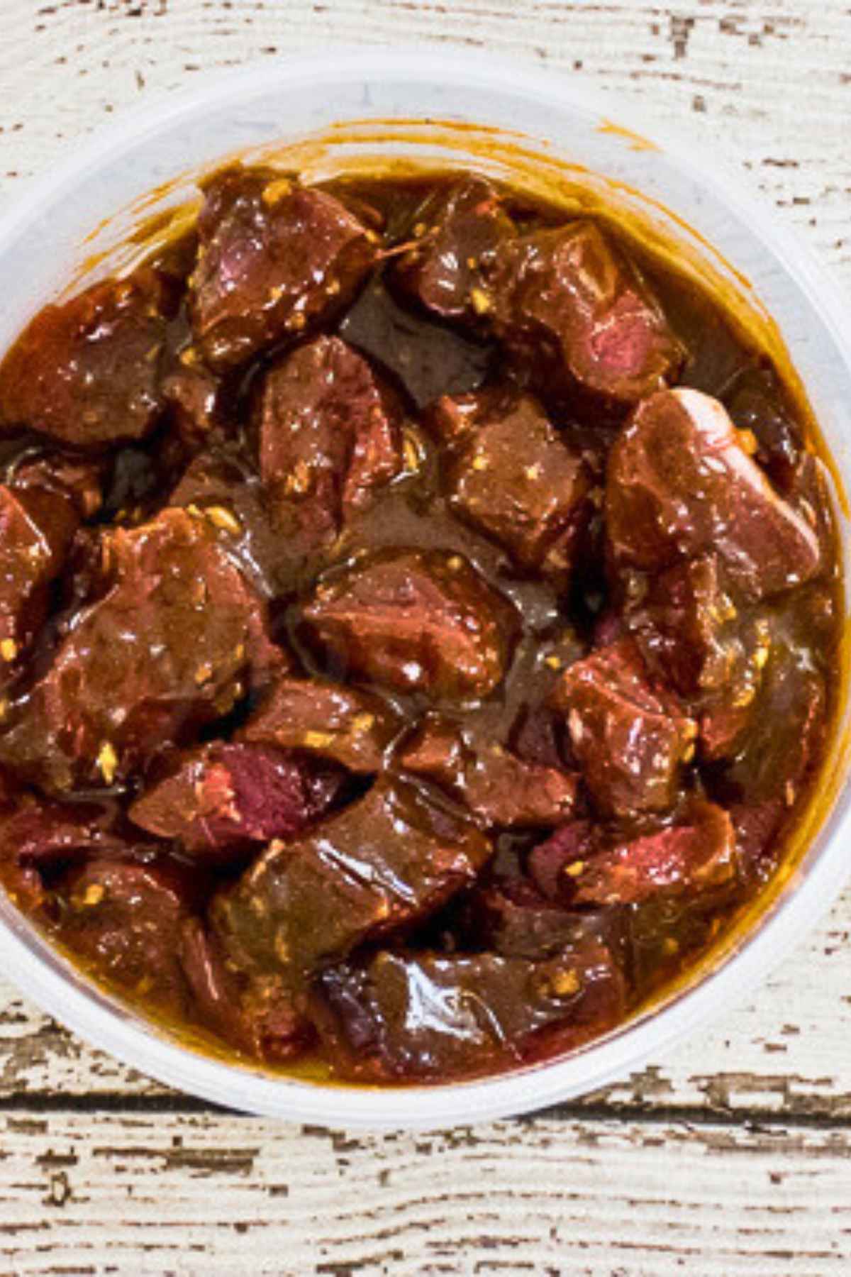 Raw venison tenderloin cubes coated in a dark marinade, sitting in a round plastic container on a rustic white surface.