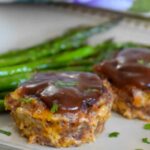 Two baked BBQ Cheddar Meatloaves served on a plate with roasted asparagus, topped with glossy barbecue sauce and garnished with chopped parsley.