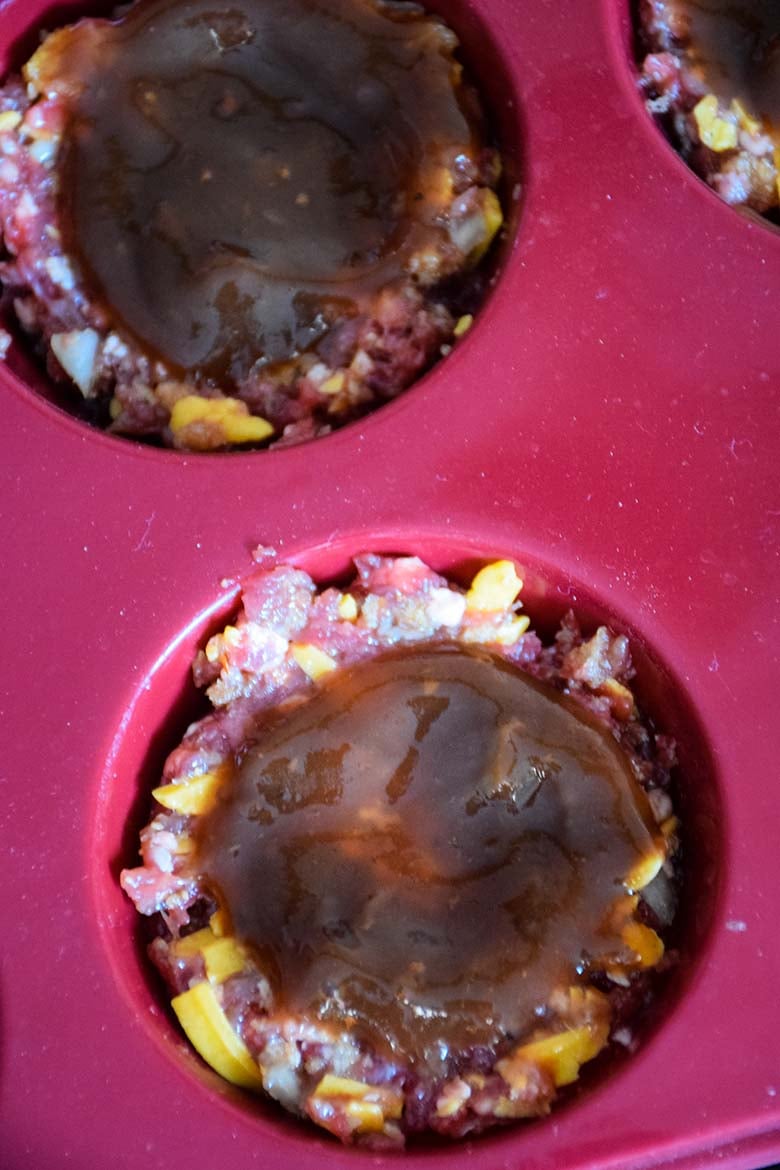 Unbaked mini meatloaves in a red silicone muffin pan, topped with a layer of barbecue sauce, showing visible bits of cheddar and onions in the raw meat mixture.