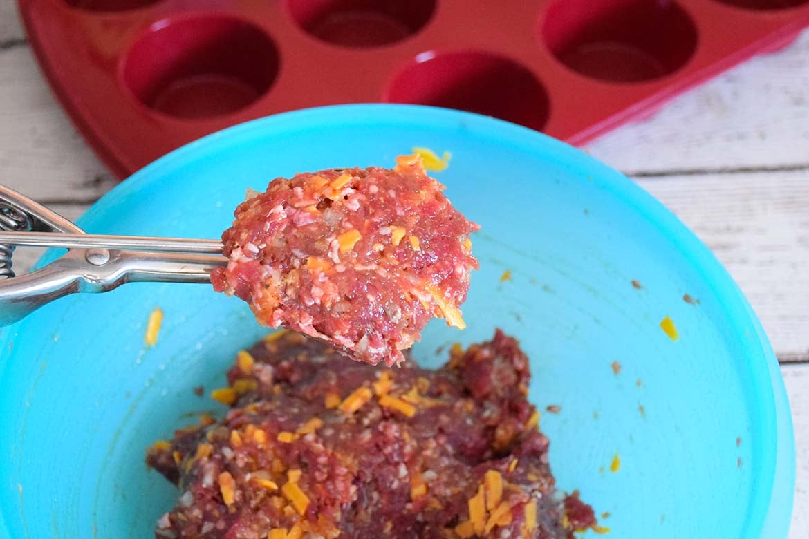 Portion of meatloaf mixture scooped from a blue bowl using a metal cookie scoop, with a red silicone muffin pan in the background.