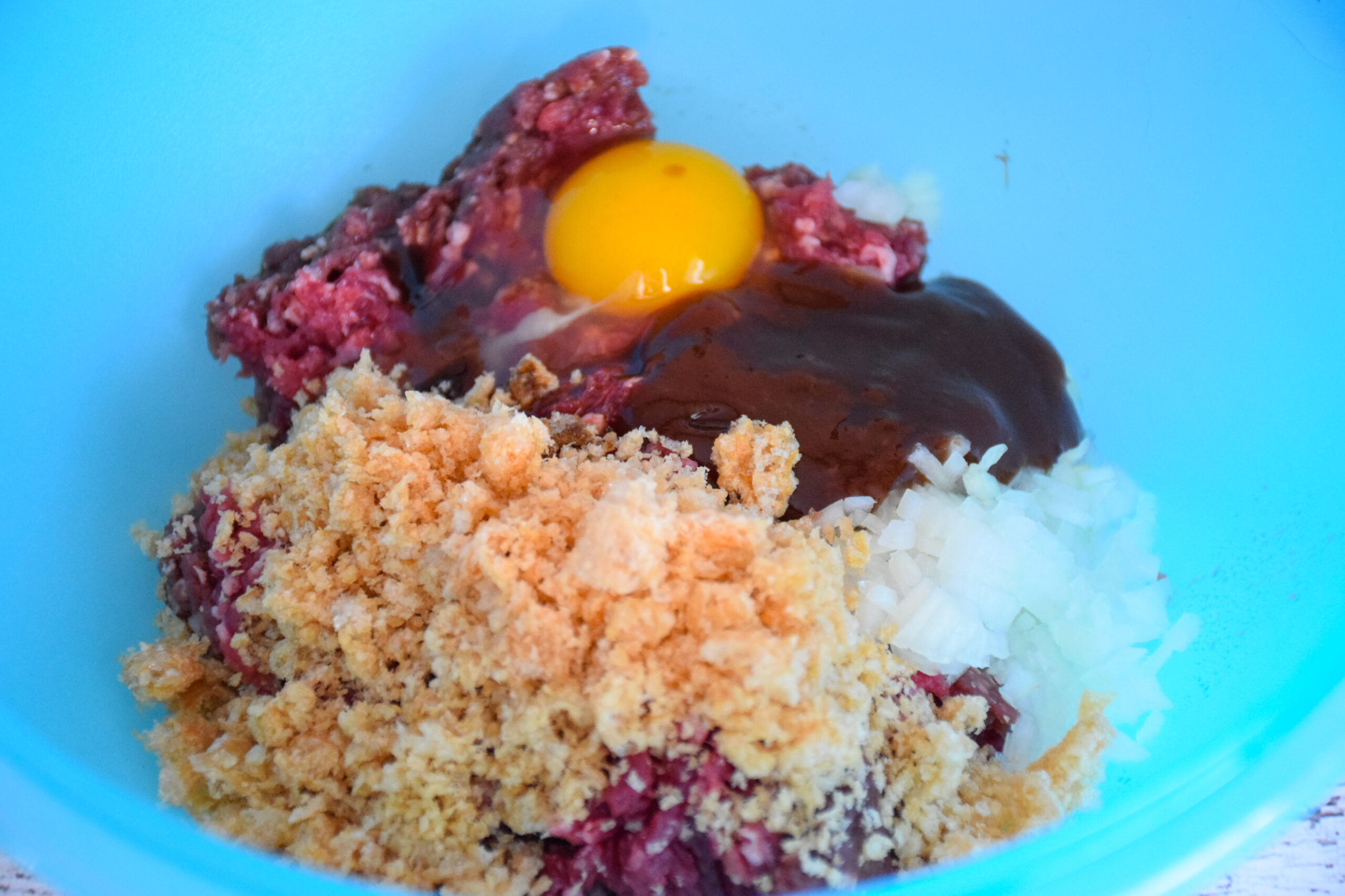 Ingredients for BBQ Cheddar Meatloaf Muffins in a blue bowl, including ground meat, a whole egg, diced onion, breadcrumbs, and barbecue sauce, ready to be mixed.