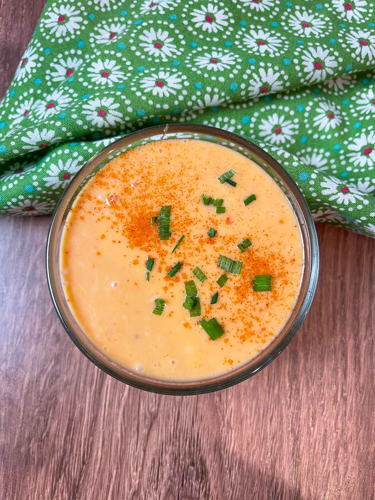 A small glass bowl of creamy orange Bang Bang Sauce garnished with chopped chives and a sprinkle of paprika, placed on a wooden surface next to a green floral napkin.