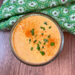 A small glass bowl of creamy orange Bang Bang Sauce garnished with chopped chives and a sprinkle of paprika, placed on a wooden surface next to a green floral napkin.