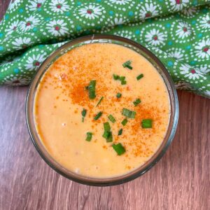 A small glass bowl of creamy orange Bang Bang Sauce garnished with chopped chives and a sprinkle of paprika, placed on a wooden surface next to a green floral napkin.