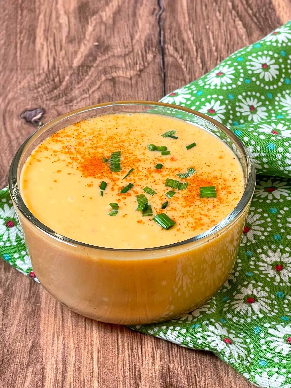 A small glass bowl of creamy orange Bang Bang Sauce garnished with chopped chives and a sprinkle of paprika, place don a wooden surface next to a green floral napkin.