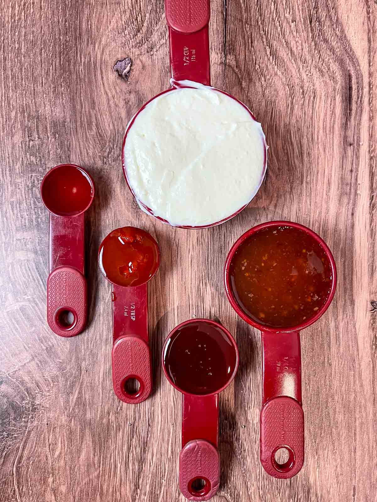 Red measuring cups filled with the following ingredients: mayonnaise, thai sweet chili sauce, sriracha, honey, and rice vinegar arranged on a wooden surface.