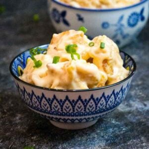 A small patterned bowl filled with crispy battered shrimp coated in a creamy sauce, garnished with chopped green onions, with another similar bowl blurred in the background.