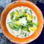A bowl of creamy green bean soup with chunks of potato, topped with shaved cheese and herbs, served in a rustic orange bowl.