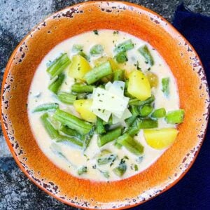 A bowl of creamy green bean soup with chunks of potato, topped with shaved cheese and herbs, served in a rustic orange bowl.