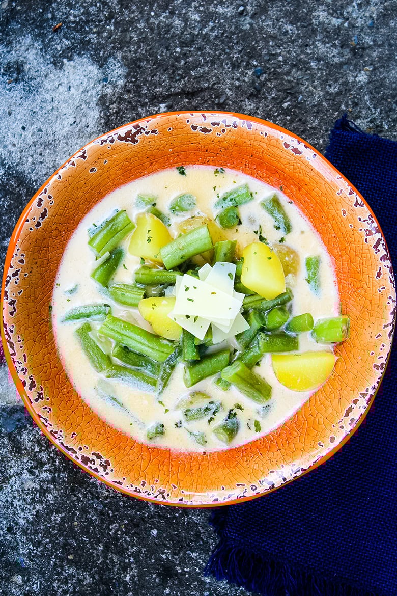 A bowl of creamy green bean soup with chunks of potato, topped with shaved cheese and herbs, served in a rustic orange bowl.
