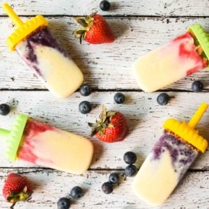 Overhead shot of berry swirled cheesecake popsicles on a white wooden surface, surrounded by fresh strawberries and blueberries.