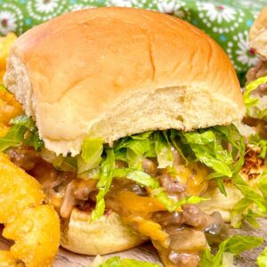 A close-up of a Big Mac sloppy joe sandwich with shredded lettuce, melted cheese, and a saucy ground beef on a soft hamburger bun served on a wooden surface with a side of golden tater tors.