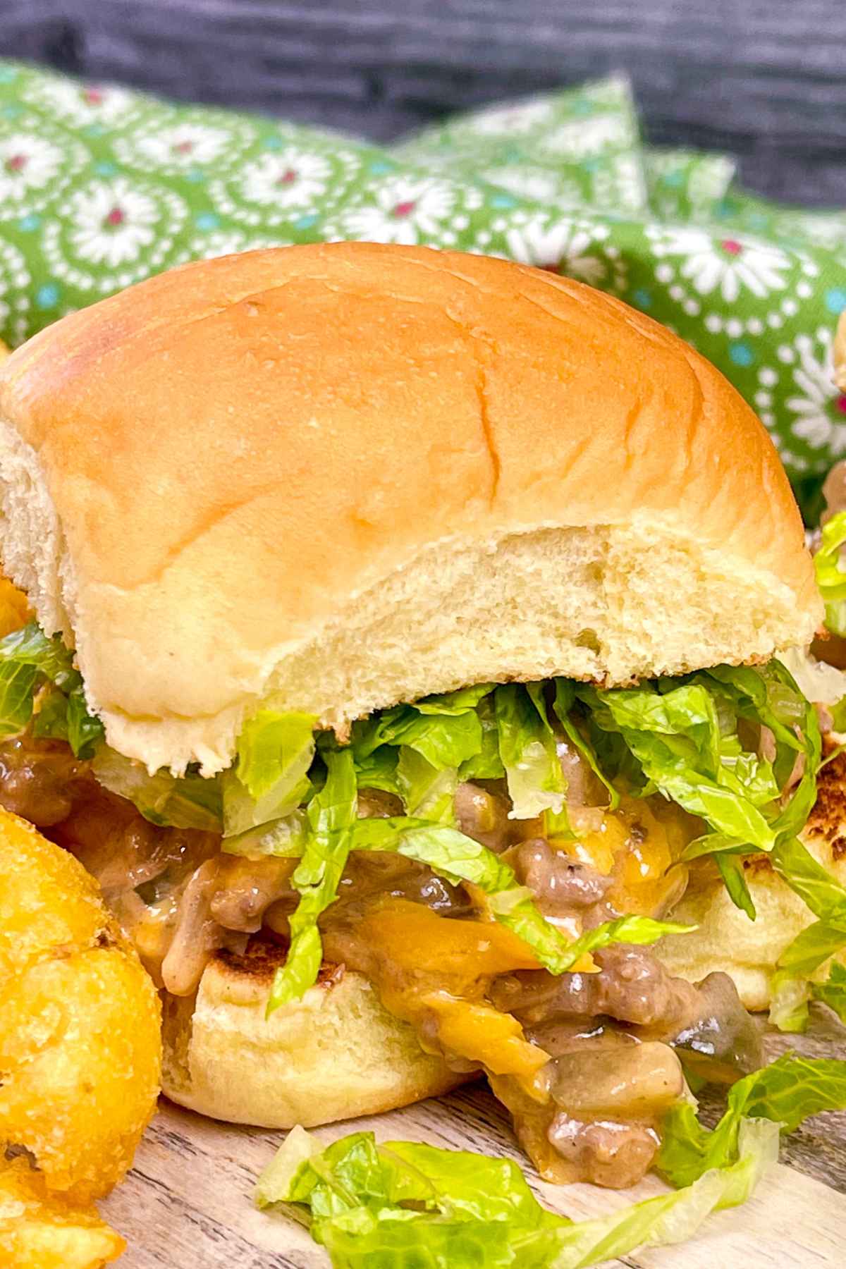 A close-up of a Big Mac sloppy joe sandwich with shredded lettuce, melted cheese, and a saucy ground beef on a soft hamburger bun served on a wooden surface with a side of golden tater tots.