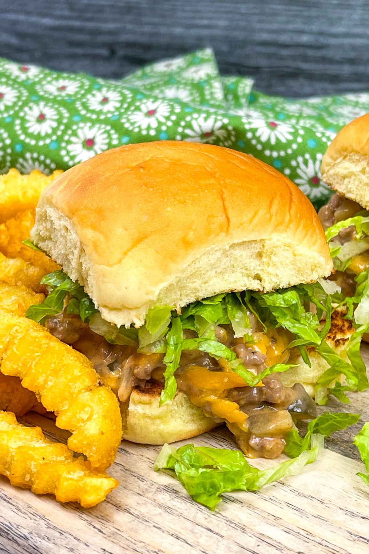 A close-up of a Big Mac sloppy joe sandwich with shredded lettuce, melted cheese, and a saucy ground beef on a soft hamburger bun served on a wooden surface with a side of golden tater tots.