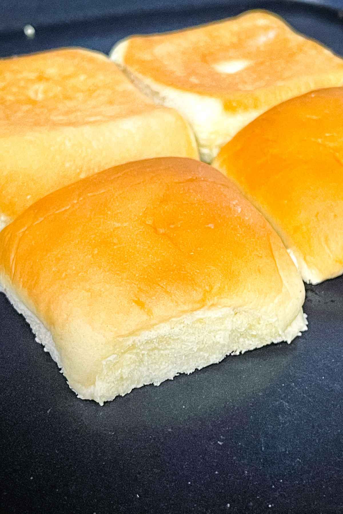 Four sandwich buns being toasted on a griddle, with golden-brown tops and soft, fluffy edges visible.