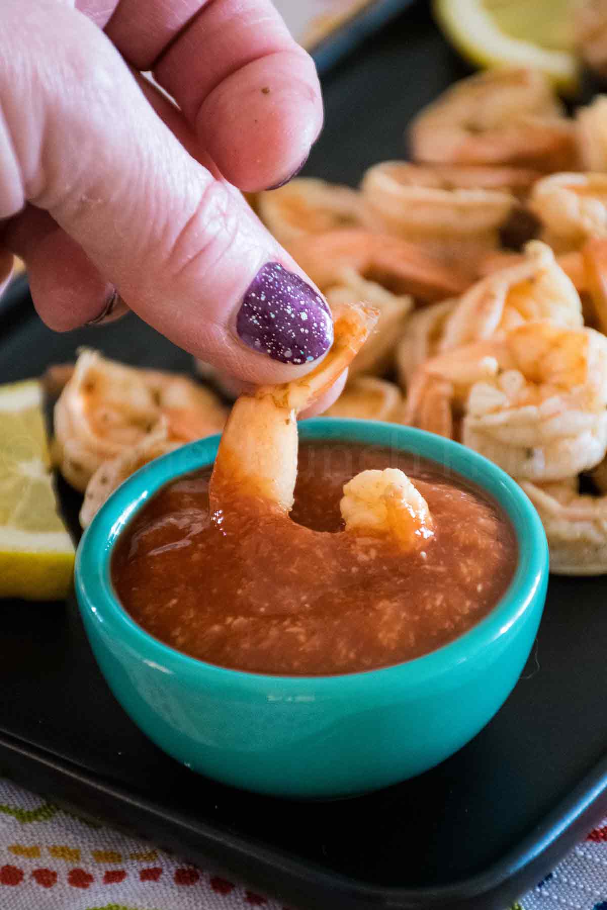Hand with purple glittery nail polish dipping a cooked shrimp into a bowl of cocktail sauce with a plate of shrimp and lemon slices in the background.