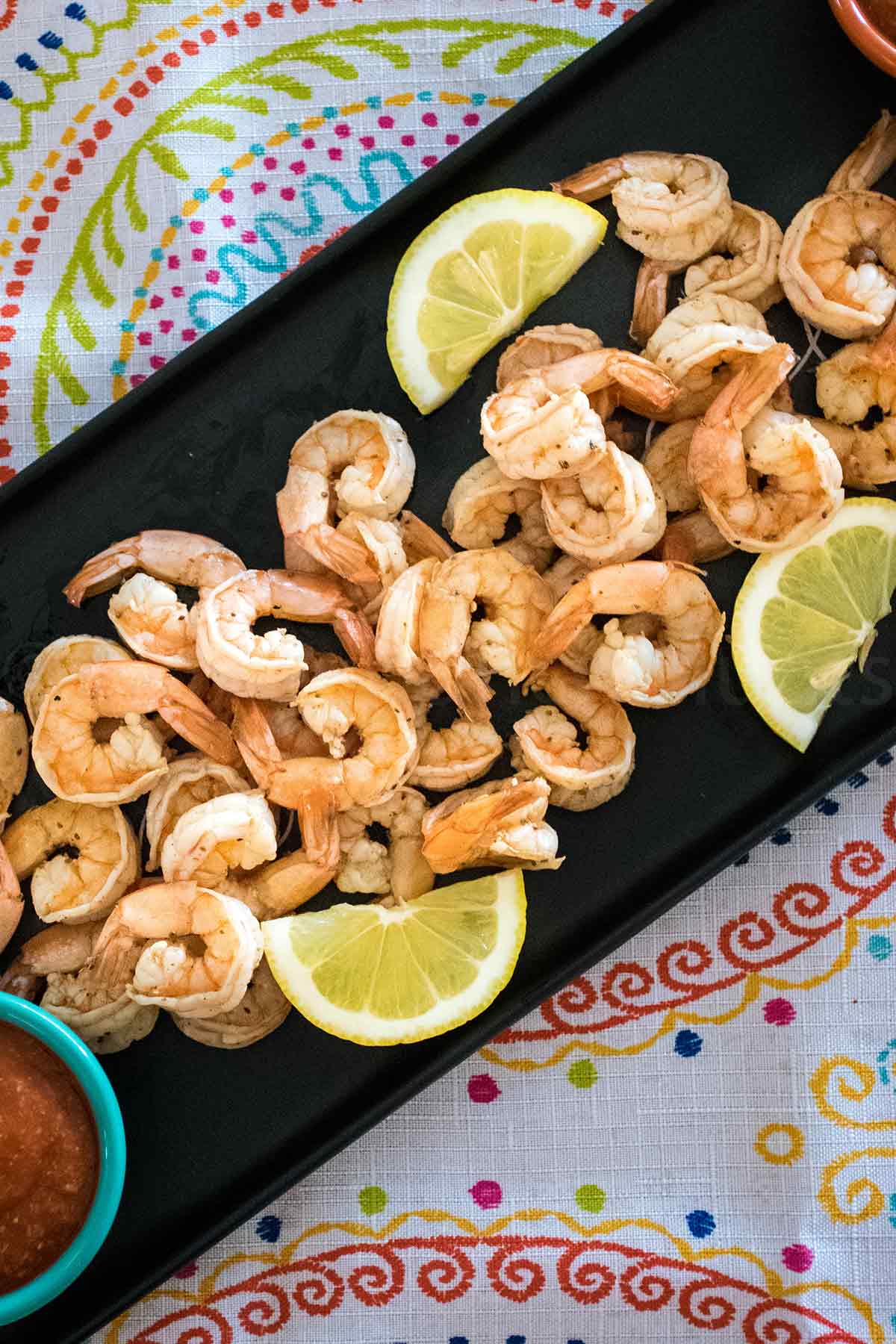 Overhead view of a platter of cooked peeled shrimp garnished with lemon wedges, served on a dark tray with a colorful napkin underneath.