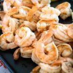 Side view of a platter of cooked peeled shrimp garnished with lemon wedges, served on a dark tray with a colorful napkin underneath.