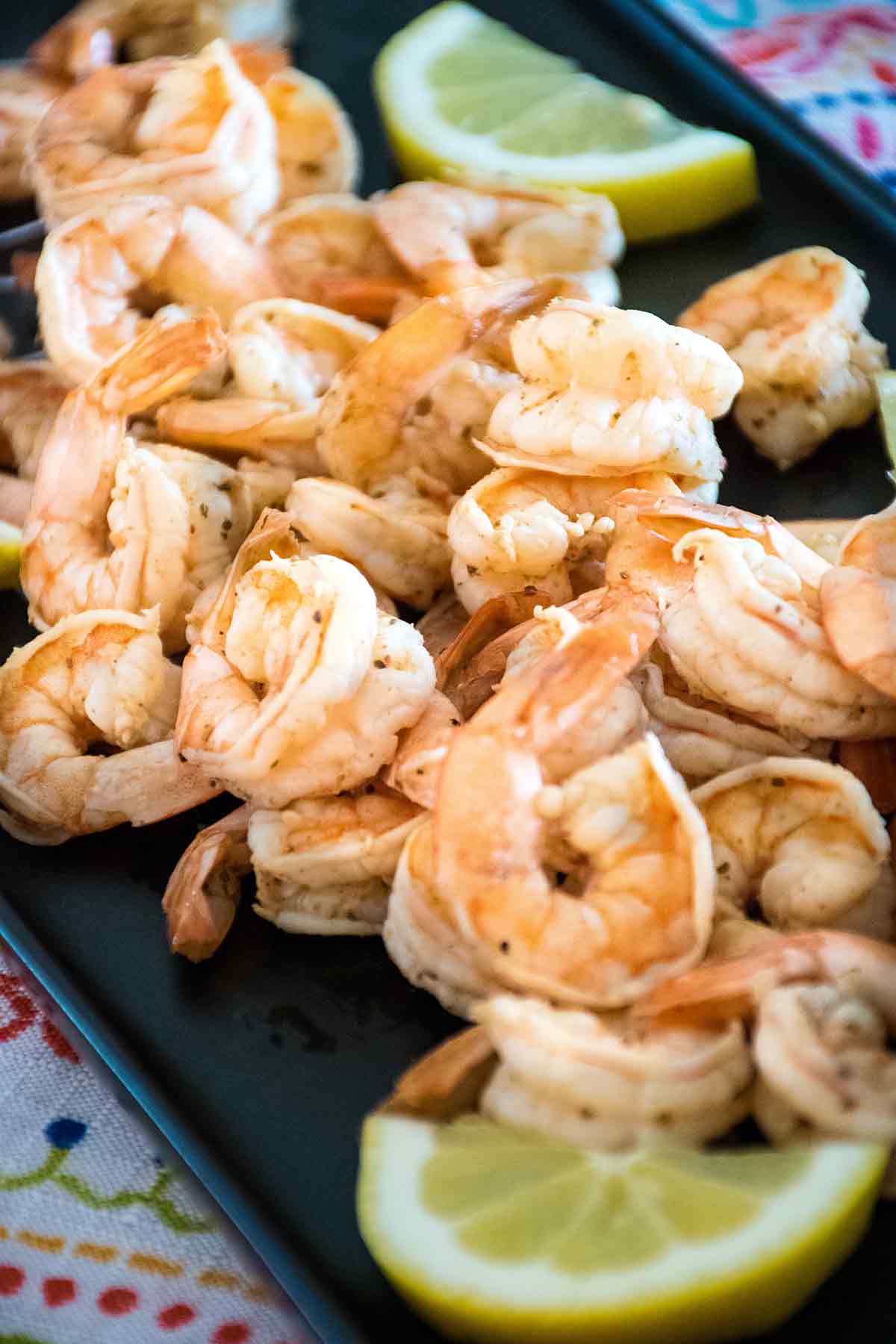 Side view of a platter of cooked peeled shrimp garnished with lemon wedges, served on a dark tray with a colorful napkin underneath.