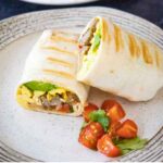 Two halves of a grilled breakfast burrito on a plate, served with cherry tomato halves and a sprig of fresh cilantro.