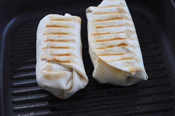 Two burritos with grill marks cooking in a ridged grill pan, ligihtly browned on the bottom side.