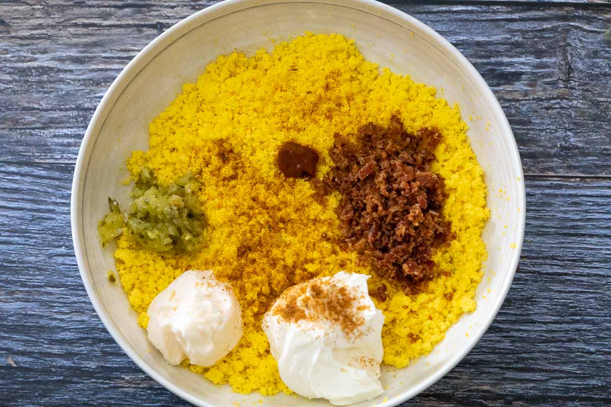 Bowl of deviled egg yolk mixture ingredients, including crumbled bacon, mayonnaise, sour cream, relish, hot sauce, and spices on top of bright yellow mashed egg yolks.