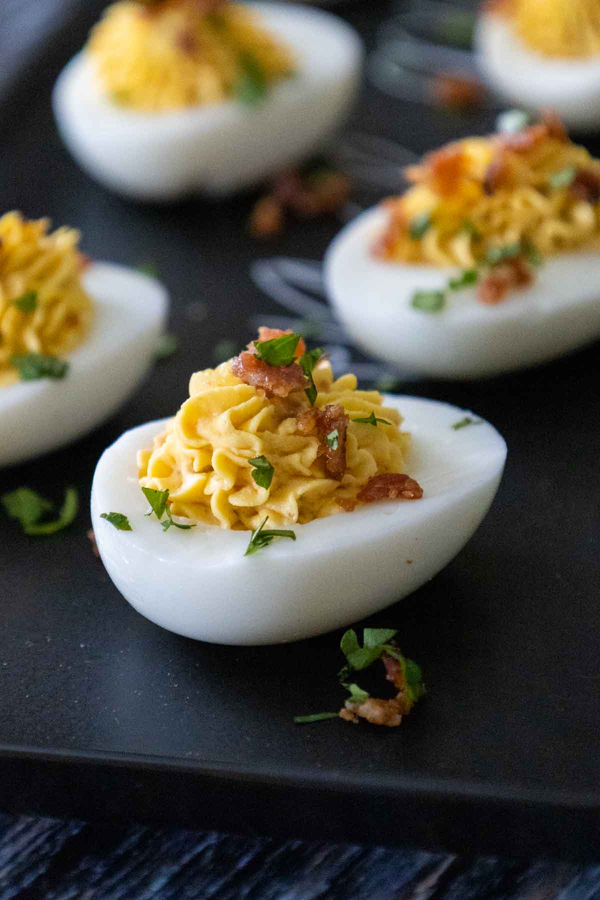Deviled Egg on a black plate, filled with piped yellow yolk mixture, garnished with bacon crumbles and fresh herbs.