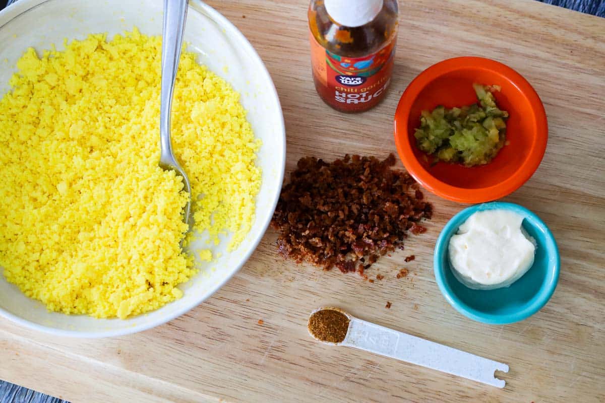 Ingredients for deviled egg filling arranged on a wooden board next to board next to a bowl of mashed egg yolks, including bacon bits, relish, mayonnaise, hot sauce, and cajun seasoning.