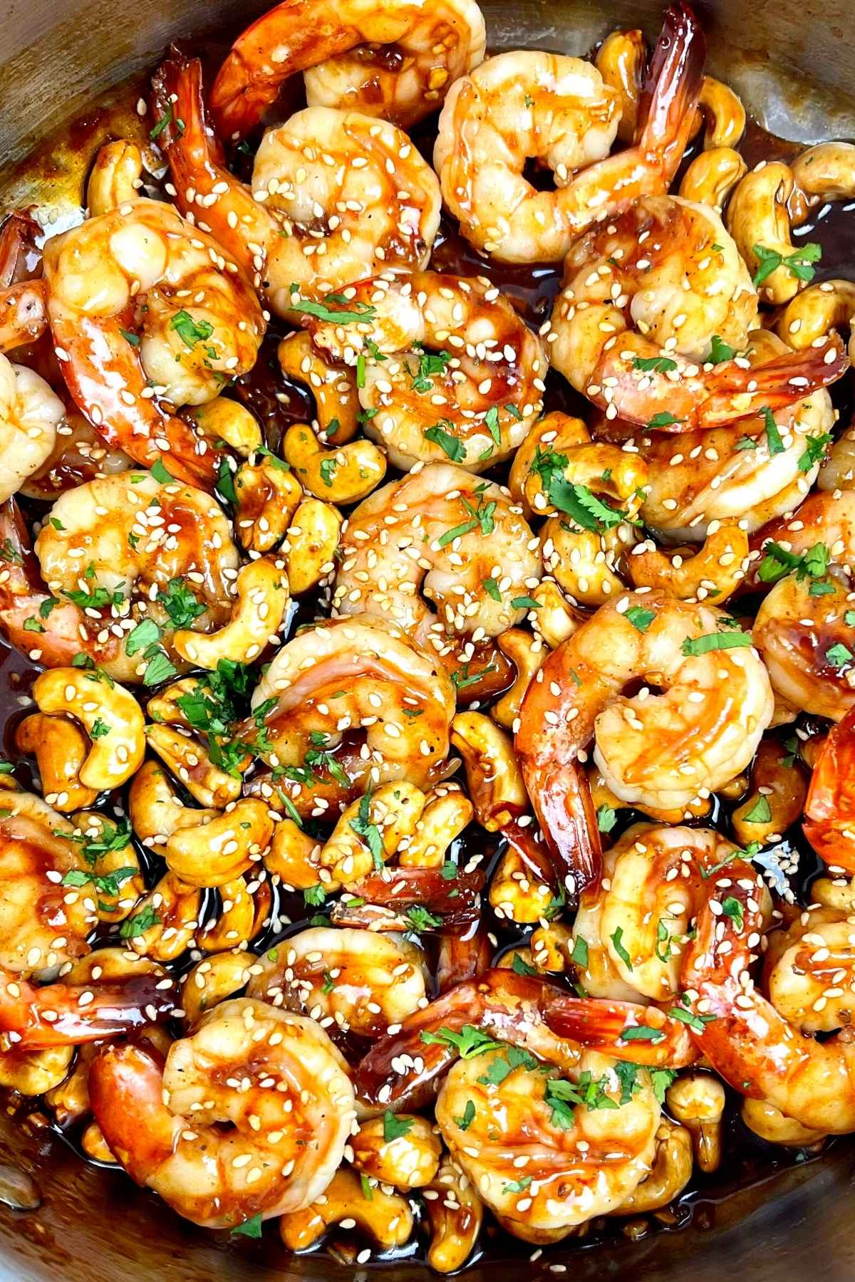 Cashew shrimp in a skillet coated in glossy sauce and sprinkled with sesame seeds and chopped herbs.