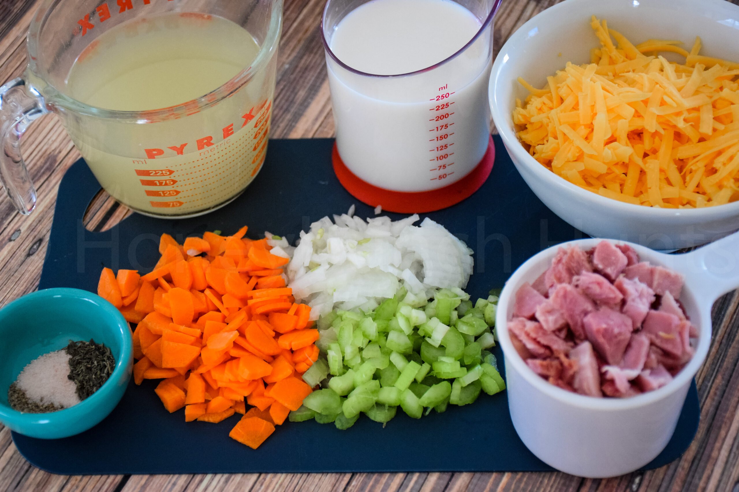 Chopped ingredients for a casserole recipe arranged on a cutting board: diced carrots, celery, onions, and ham; shredded cheddar cheese in a bowl; mlk and broth in measuring cups: and a small bowl of salt and herbs.