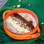Orange bowl of chunky monkey noatmeal topped with white chocolate chips, chopped pecans, and melted chocolate, set on a green cloth over a rustic wood table.
