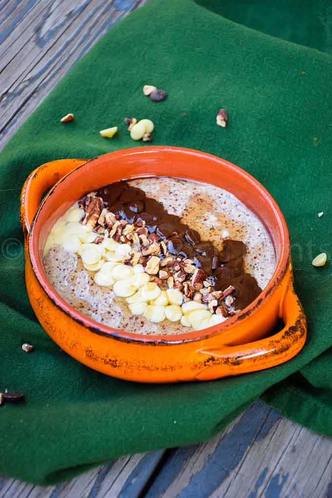 Orange bowl of chunky monkey noatmeal topped with white chocolate chips, chopped pecans, and melted chocolate, set on a green cloth over a rustic wood table.