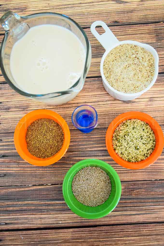 Top down view of ingredients for no grain cereal, including a glass measuring cup of milk, a white measuring cup of almond flour, and a small colorful bowl of flaxseed meal, hemp seed, chia seed, and a small measuring glass of extract, all arranged on a wooden surface.