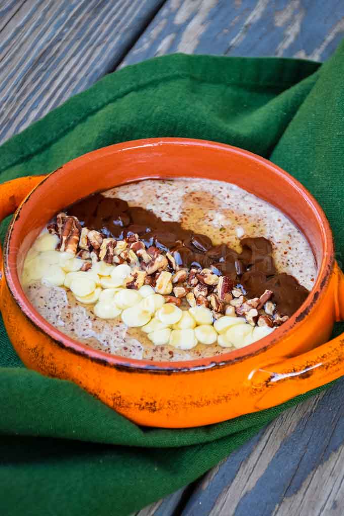 Orange bowl of chunky monkey noatmeal topped with white chocolate chips, chopped pecans, and melted chocolate, set on a green cloth over a rustic wood table.