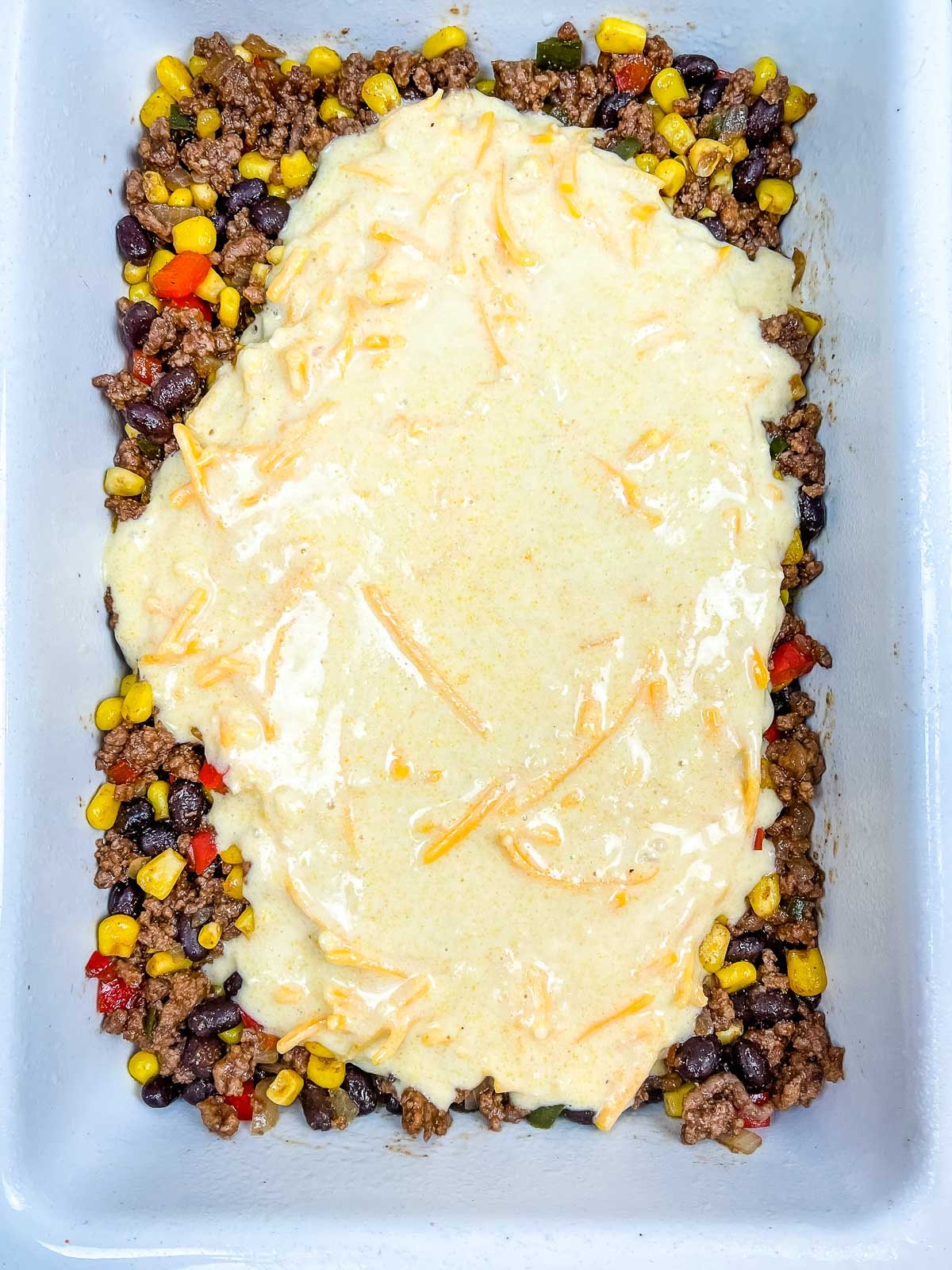 Casserole dish with a cooked beef, corn and bean base, partially topped with cornbread batter before baking.