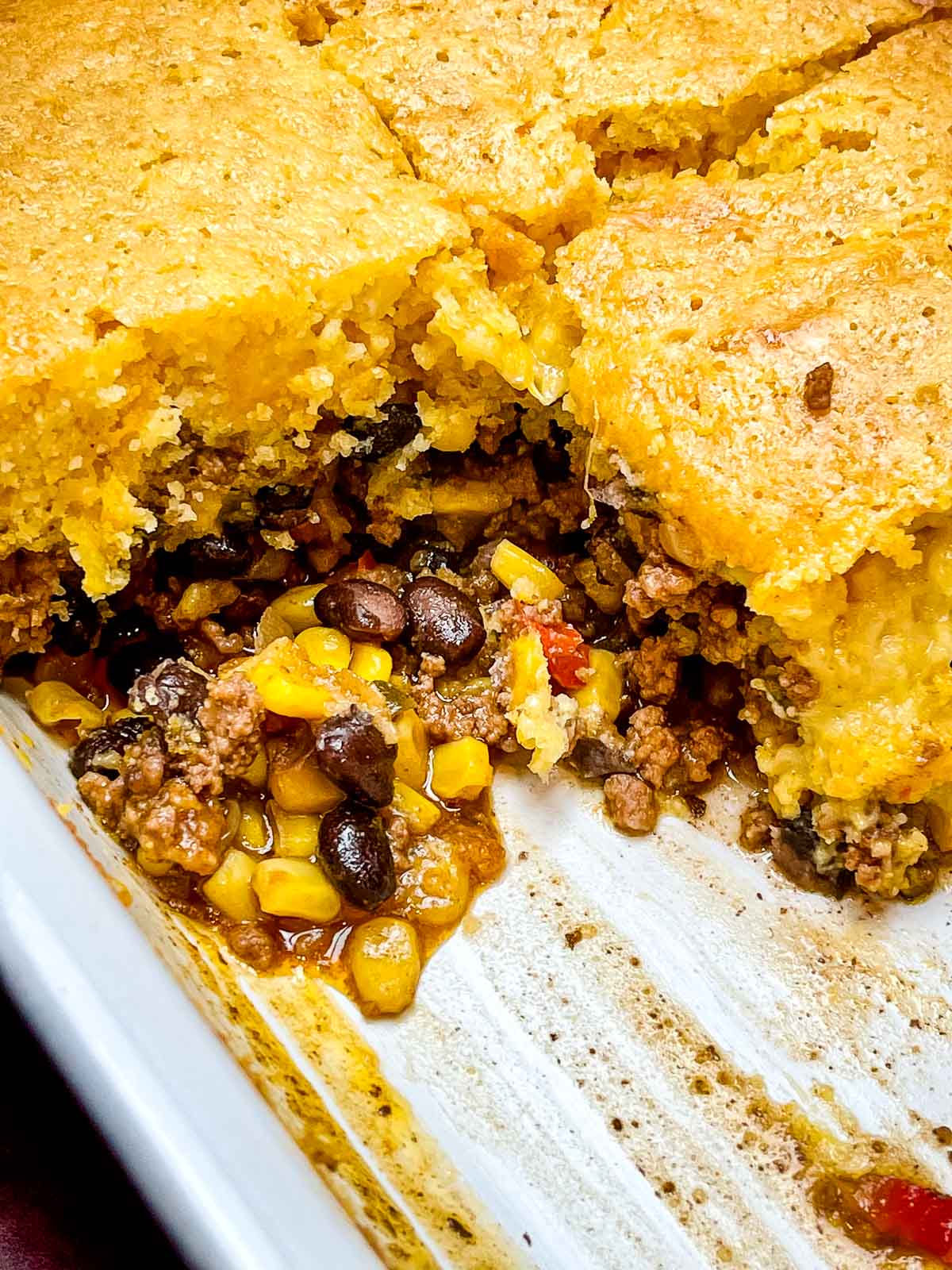 Close up of sliced cowboy cornbread casserole in a baking dish, showing layers of golden cornbread crust and a juicy beef, corn, and black bean filling.