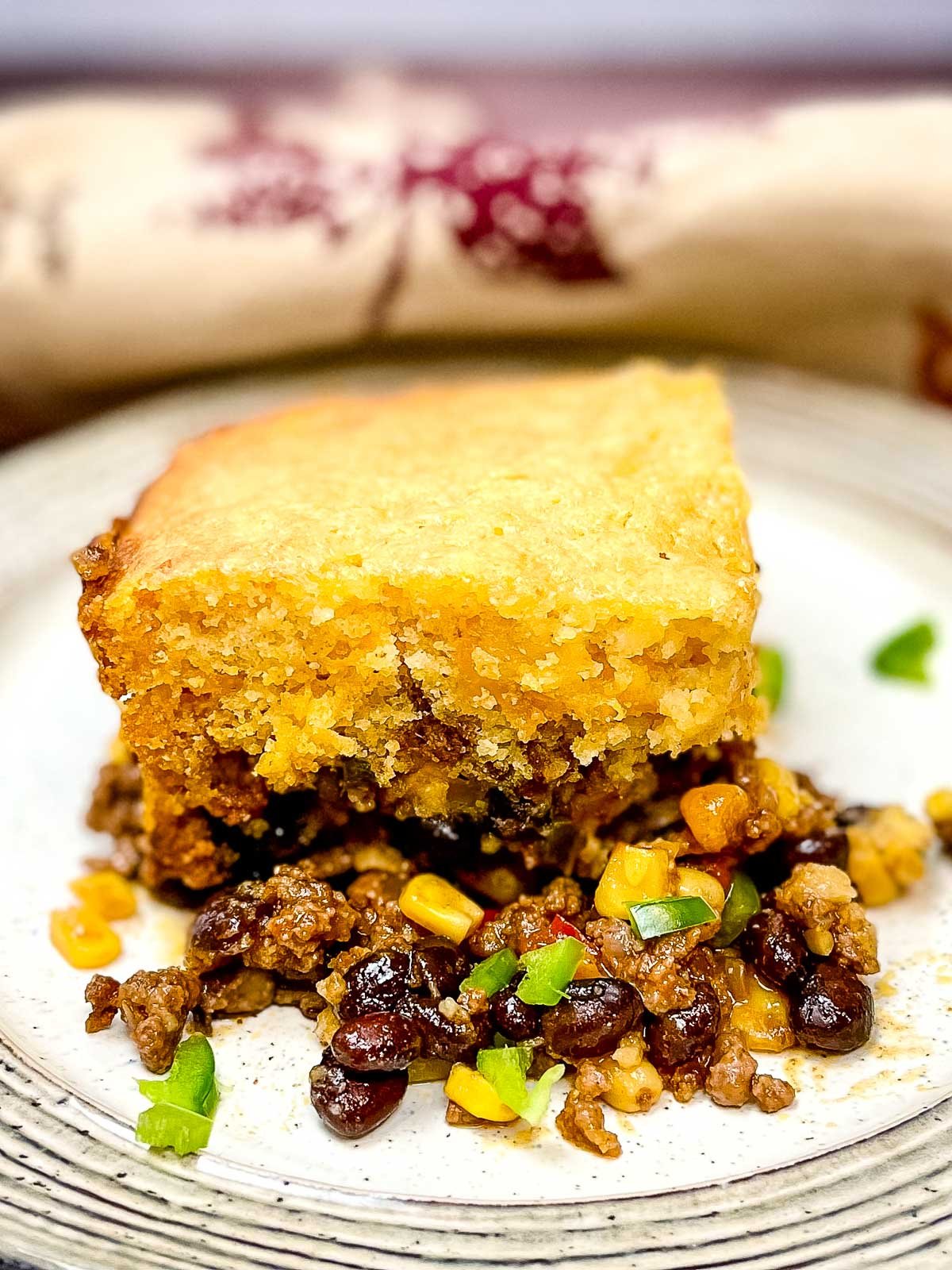 A hearty serving of cowbowy casserole on a white plate, featuring a thick golden cornbread topping over a flavorful filling of ground beef, black beans, corn, and diced vegetables.