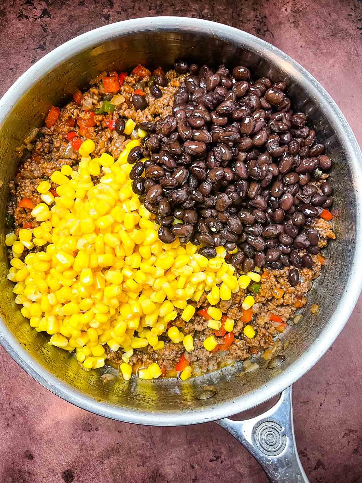 Ground beef mixture in a skillet topped with separate piles of black beans and corn, ready to be mixed into the seasoned meat base.