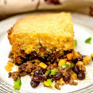 A hearty serving of cowbowy casserole on a white plate, featuring a thick golden cornbread topping over a flavorful filling of ground beef, black beans, corn, and diced vegetables.